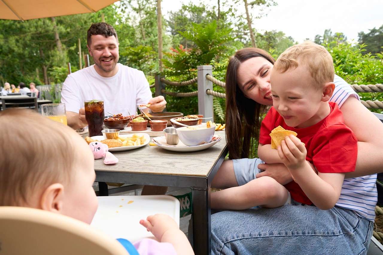 Boy eats a tortilla chip on a woman's lap; a man and baby sit at a table with fries and drinks, dining together in a leafy outdoor restaurant under umbrellas.