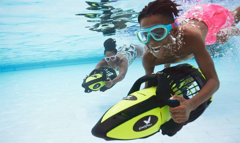 Two goggle-wearing swimmers ride handheld electric sea scooters, propelling forward underwater in a clear tiled pool; bubbles trail behind. The scooters show an oval logo with small, unreadable white text.