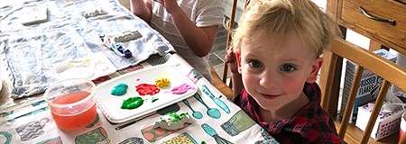 Child paints clay pieces at a dining table. Palette with bright paints and a rinsing cup nearby. Another child works beside them; wooden chairs and drawers frame the home setting.