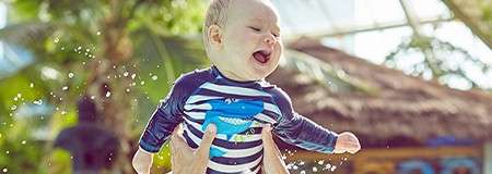 Baby laughing, lifted by hands above splashing water in a sunny tropical setting, wearing a striped long-sleeve swimsuit with blue whale graphic; palm trees and a thatched-roof building behind.