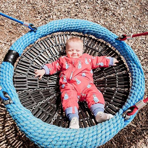 Baby lies sprawled and squinting, wearing a red onesie with blue cuffs, resting on a circular blue rope net swing held by chains, in a playground over wood-chip ground.