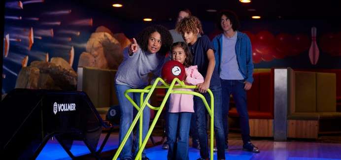 Child pushes bowling ball down a metal ramp while adults guide and watch in a bowling alley. "6" on the red ball; "VOLLMER" on the black machine.