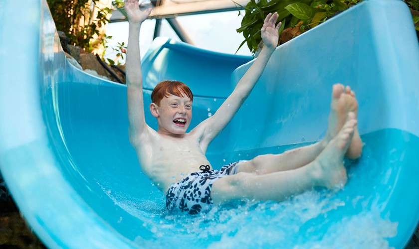 Child slides down a bright blue waterslide, arms raised and smiling, feet forward. Water splashes around. Surroundings include indoor structures and green plants along the slide.