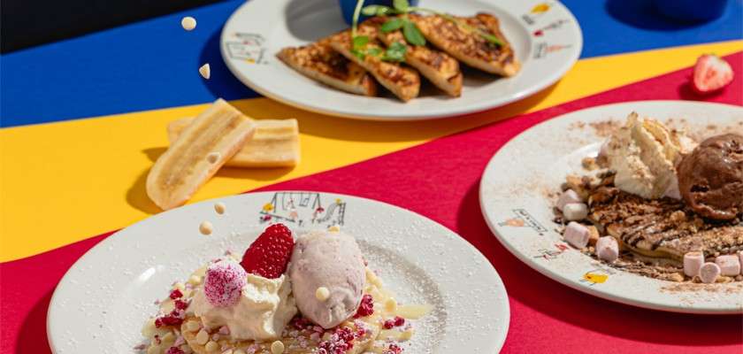 Dessert plates sit arranged, topped with ice cream scoops, whipped cream, berries, and marshmallows; white chocolate pearls fall midair. Context: colorful red, yellow, and blue tabletop with additional sweet dishes.