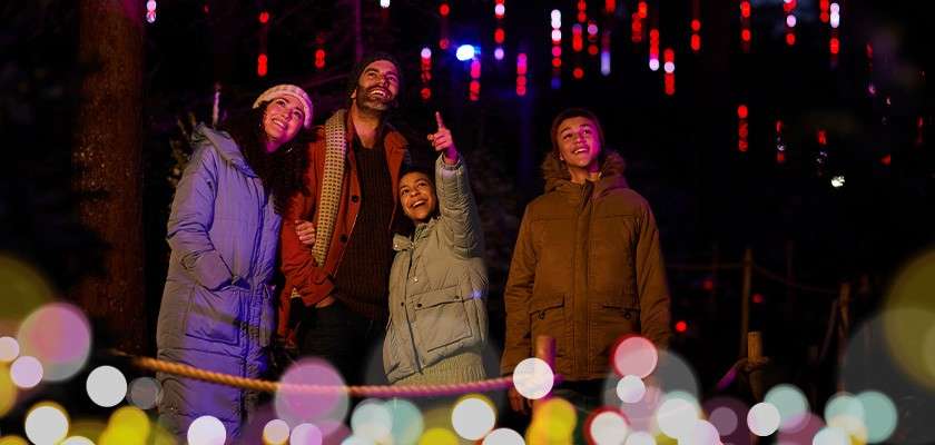 Four bundled-up people stand together, one child pointing upward, as they admire hanging red lights. Context: nighttime outdoor path in a forest, rope barrier and colorful bokeh foreground.