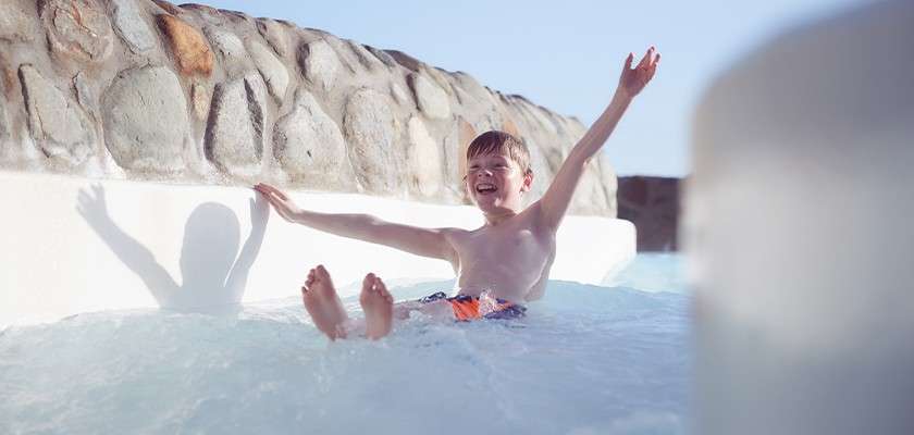 Boy slides down a water slide, arms raised and smiling, feet forward. Sunlit water splashes around him, with a white flume, stone wall, and blue sky in the background.
