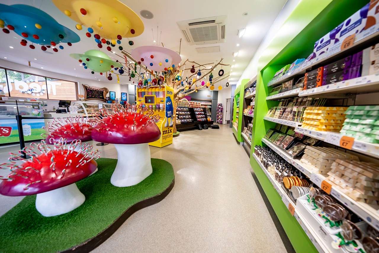 Candy shop shelves display chocolates while whimsical mushroom stands hold candy canes; a yellow M&M station operates beneath colorful hanging discs. Text: "m&m"; "mix together"; "£7".
