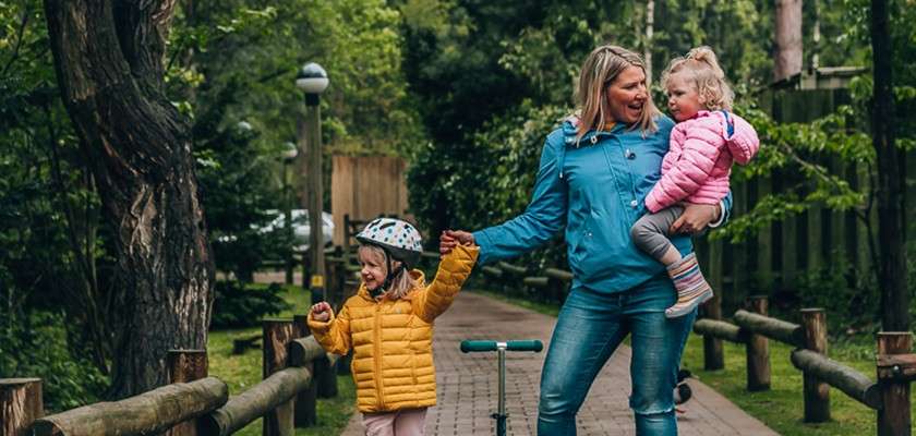 Parent carries a toddler and holds hands with an older child, walking along a tree-lined path with wooden rails; a scooter rests nearby. All wear jackets; child wears helmet.