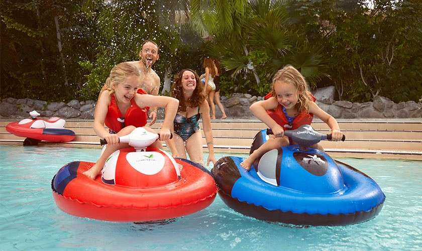 Two children ride inflatable bumper boats, steering and splashing. Behind them, two adults wade and laugh. The pool is shallow with steps, spray of water, and tropical greenery surrounding.