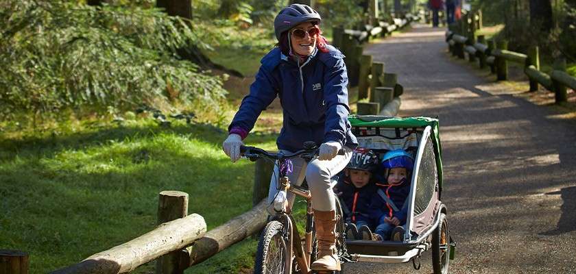 A mother cycling through the forest whilst pulling her children behind her in a carrier.
