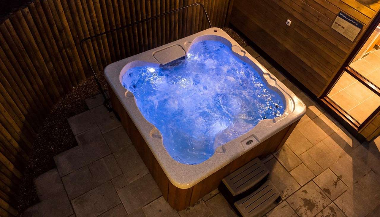 Hot tub bubbles vigorously, glowing blue, in an outdoor patio enclosure with wood-paneled walls, paver floor, a handrail, and small plastic steps at the side.