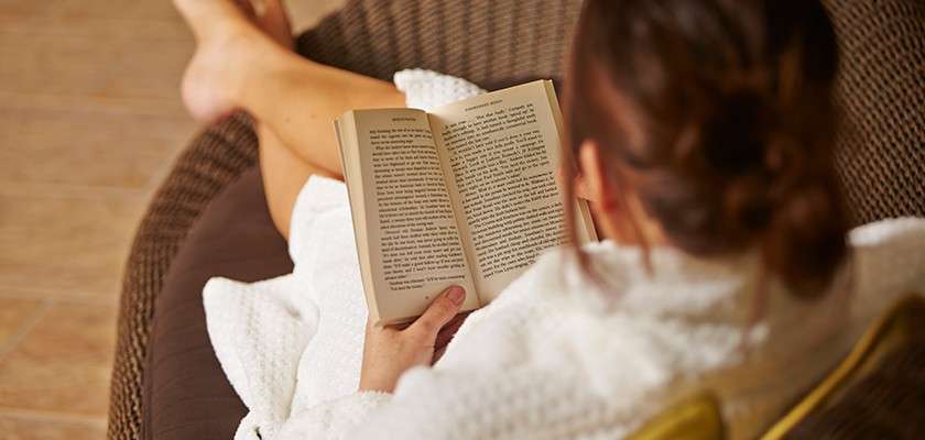 A person in a white bathrobe reads an open paperback, reclining with legs crossed on a woven lounge chair in a softly lit indoor space with a tiled floor.
