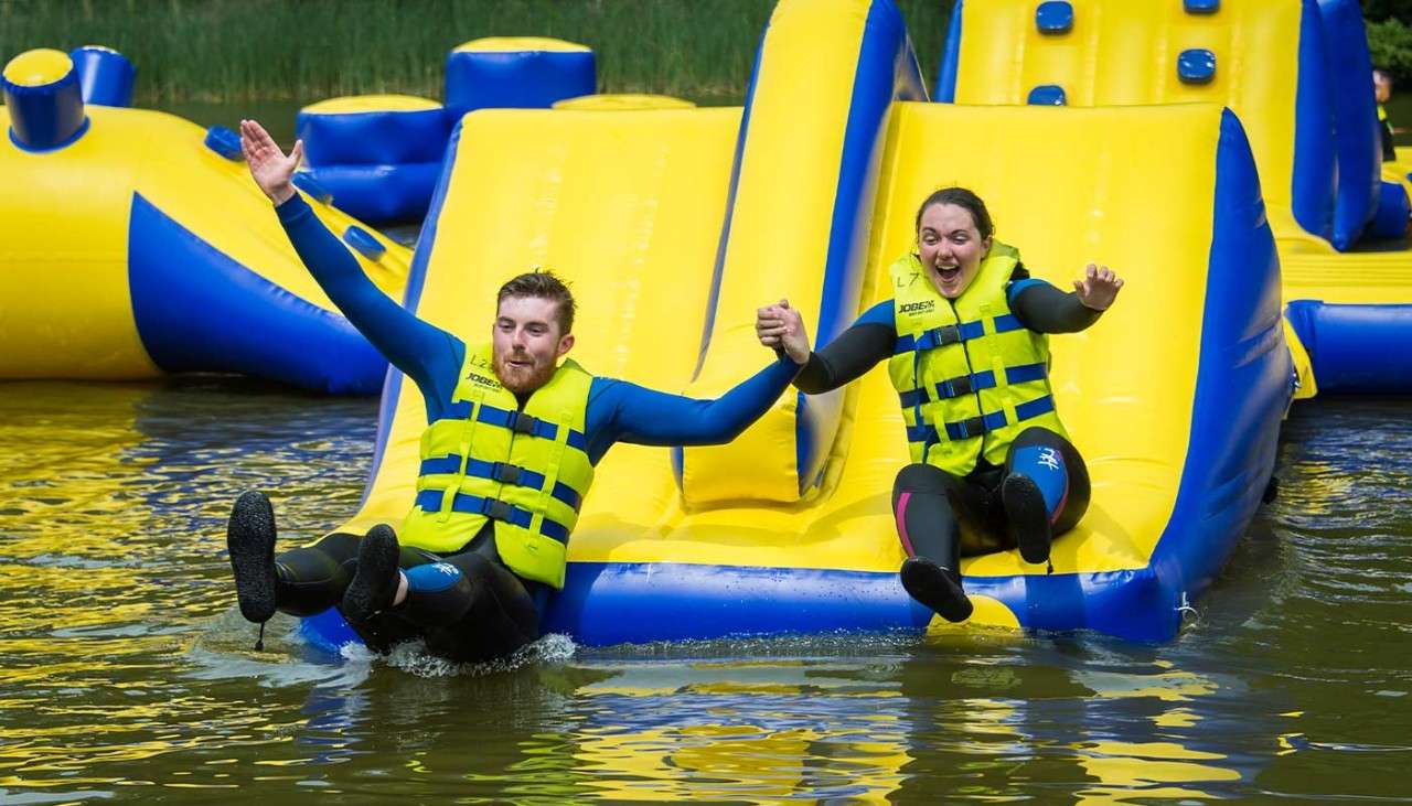 Couple sliding down Aqua Parc inflatable obstacle course on the lake