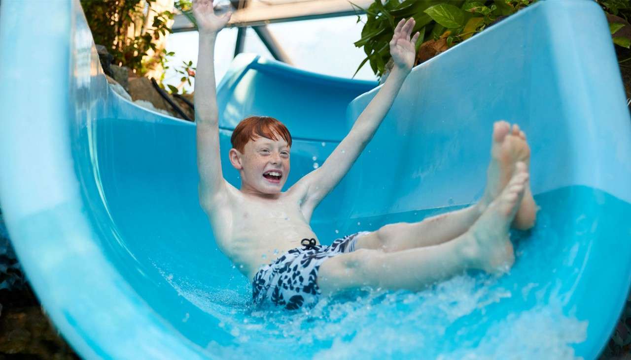 Boy sliding down a blue water slide, smiling with arms raised and feet forward. Context: splashing water in an indoor water park with plants and visible structural beams.