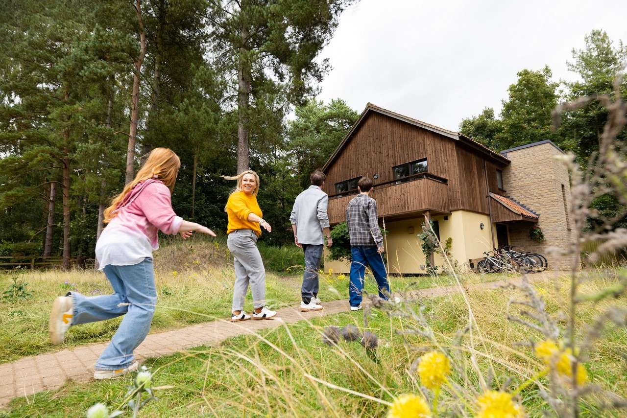 Family of four walks up a path, one turning back smiling, another running. They approach a modern wooden cabin with balcony. Bicycles rest by the entrance amid tall grass and pine trees.