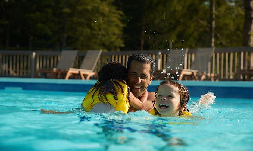 Man plays with two children wearing yellow life vests, splashing and swimming together in a clear blue outdoor pool; lounge chairs and trees line the background under daylight.