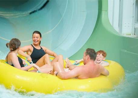 Four people ride a yellow inflatable raft, laughing, as they exit a large enclosed water slide into a splash pool at an indoor water park.