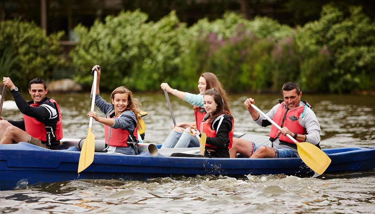 Five people in life jackets paddle a blue canoe, smiling and rowing with yellow paddles; water splashes around them as they move across a calm river bordered by green shrubs.