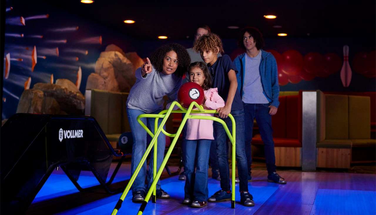 Child pushes a red bowling ball down a neon-green ramp; an adult points while family observes, in a bowling alley with benches and murals. Visible text: VOLLMER; 4; 9.