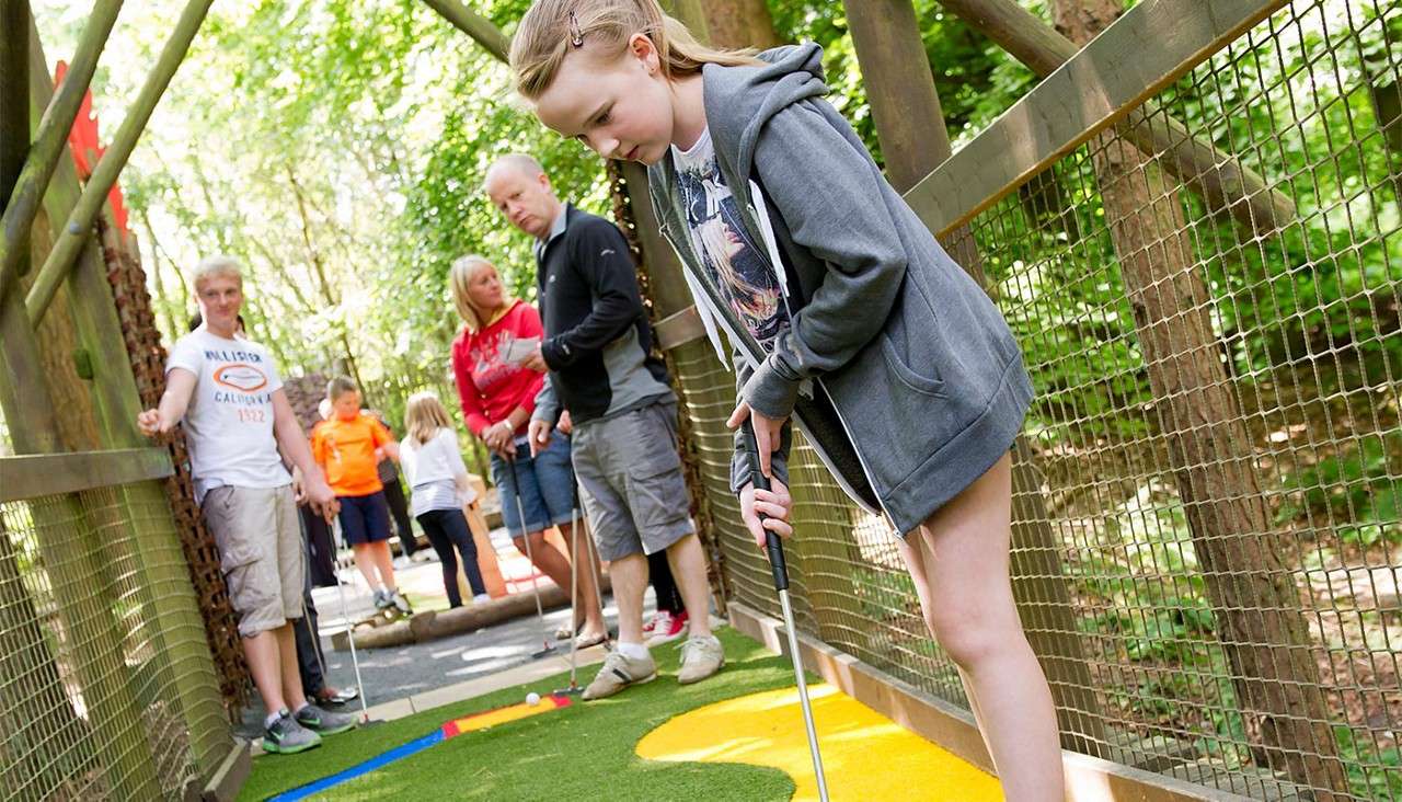 Child putts a ball on a colorful mini-golf course, leaning over with a putter; several adults and children watch behind within a wooded, netted walkway. Text: “HOLLISTER CALIFORNIA 1922”.
