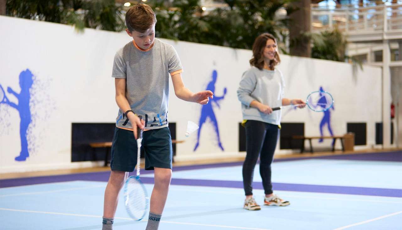 Boy tapping a shuttlecock with a badminton racket on an indoor court; a woman behind practices; blue court lines, benches, and player silhouettes on the wall. Text: VANS.