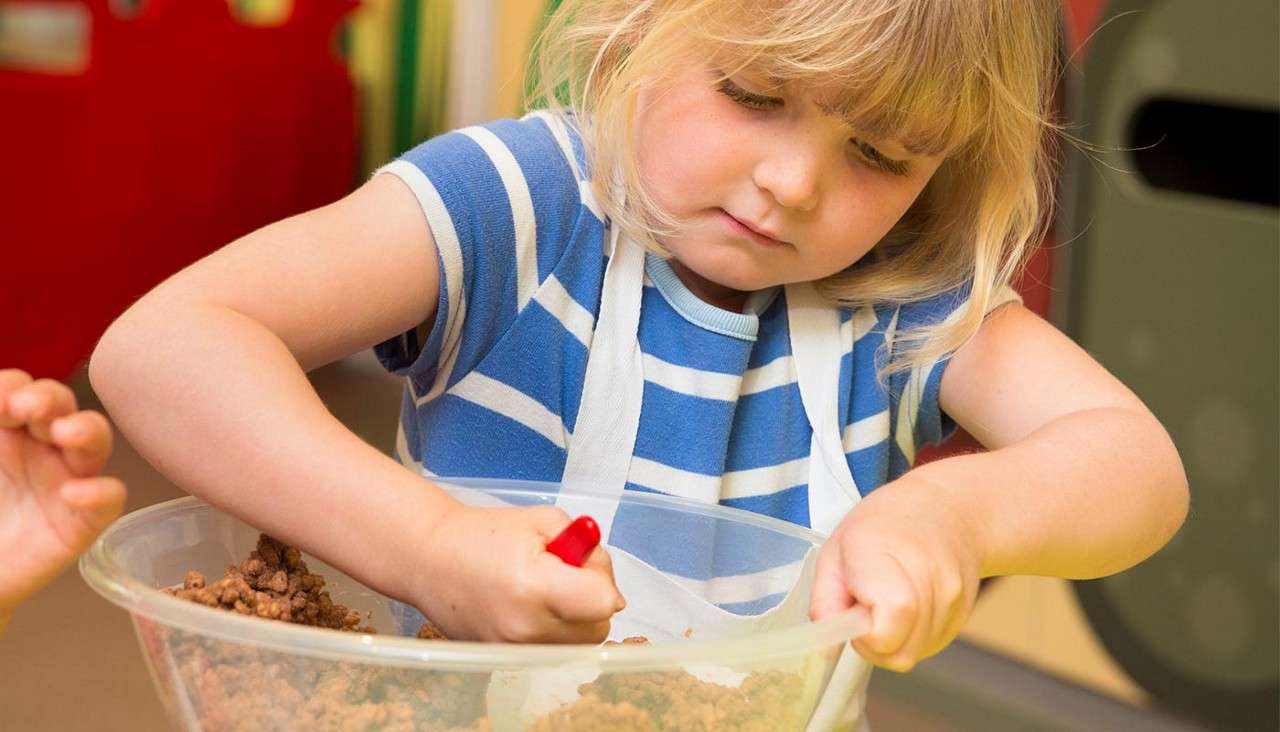 Child in striped shirt stirs crumbly mixture with red-handled utensil in a clear bowl. Context: indoor kitchen or classroom; focused expression, apron on, another child's hand nearby.