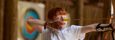 Archer draws a bow, aligning arrow near cheek, at an indoor wooden archery range with a blurred multicolored target in the background, wearing glove and arm guard, focused stance.