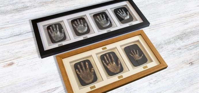 Two rectangular frames display metallic handprint casts in separate windows with small plaques beneath. One frame is black, the other wood-toned, both resting on a whitewashed wooden surface.