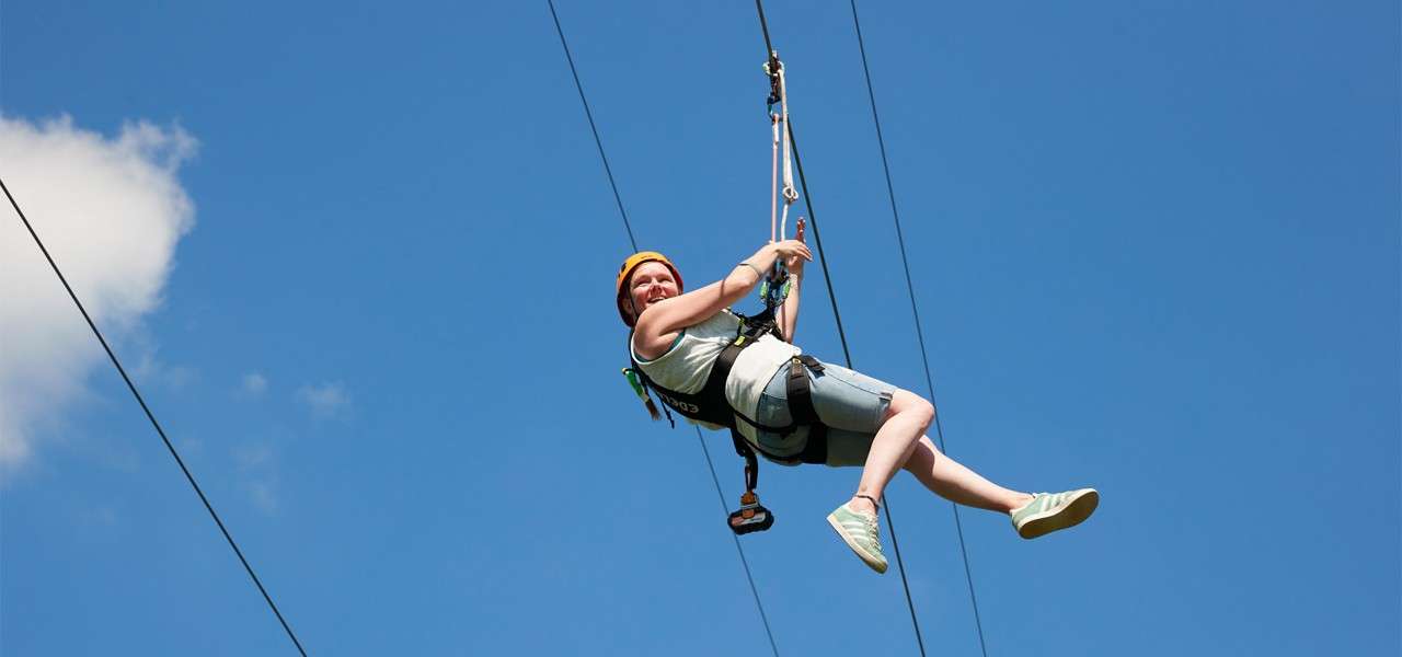 A helmeted person in a safety harness rides a zip line, gripping the rope with bent elbows, legs extended, against a clear blue sky with a small white cloud.