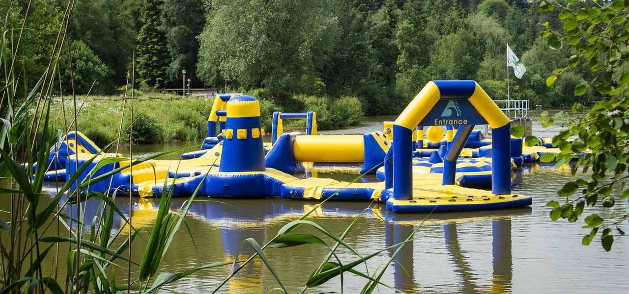 Inflatable water obstacle course floats, inviting entry through an arch labeled "Entrance", within a calm lake; yellow-and-blue towers, slides, and paths sit amid reeds and surrounding leafy trees.