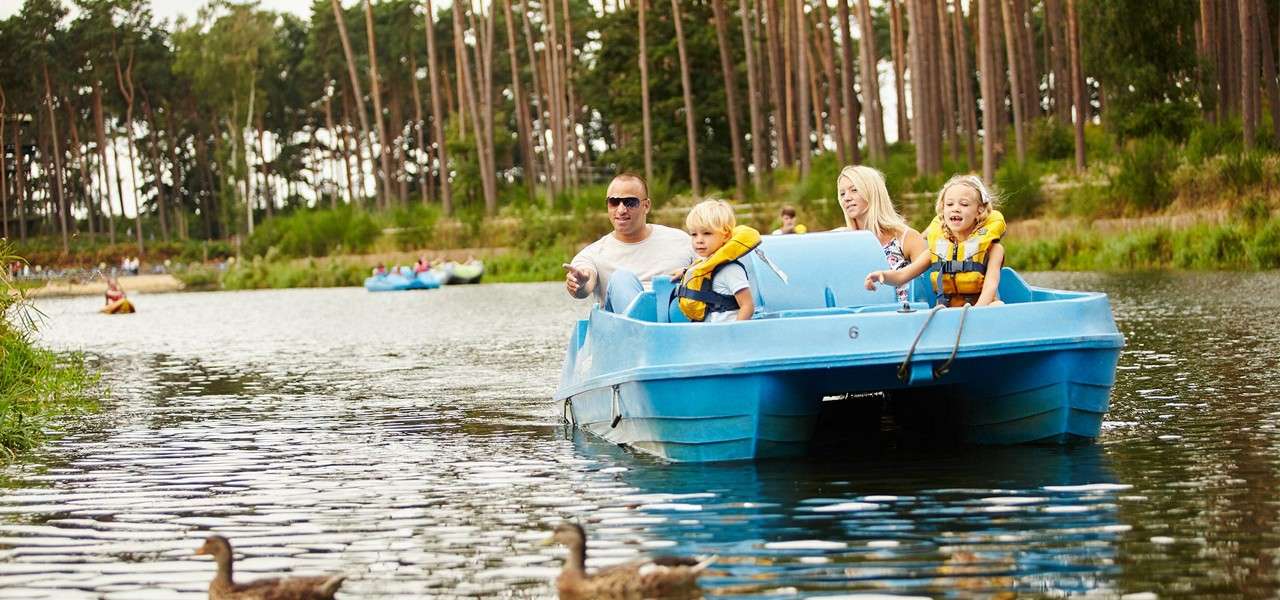 Blue pedal boat carries two adults and two children wearing life jackets, moving toward ducks. Tree-lined lake with other boats in the distance. Visible text: 6.