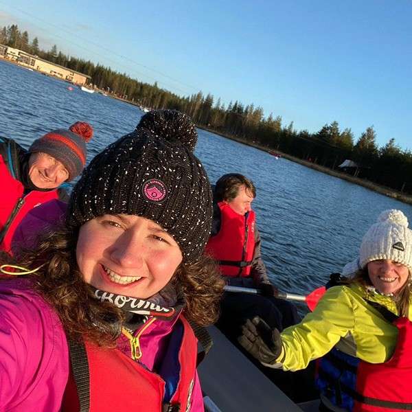 Four bundled-up people smile while boating on a calm lake. They wear red life jackets and knit hats. Conifer forest and a few lakeside buildings under a low, clear sun. Text: “ACKWOL”.