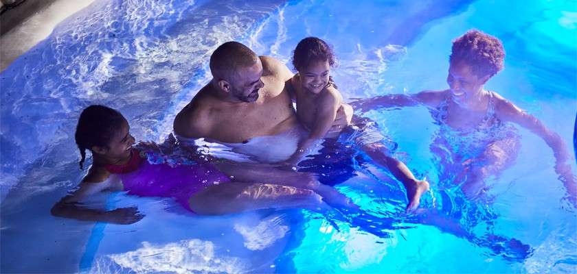 Four people splash and smile, sitting in shallow water; one adult holds a child while another reaches in. Blue pool lights glow underwater, creating a nighttime, relaxed recreational setting.