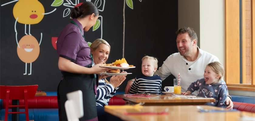 Waitress serves plates of food to a smiling family with two children at a restaurant booth, drinks on the table, wall art behind them and sunlight from large windows.