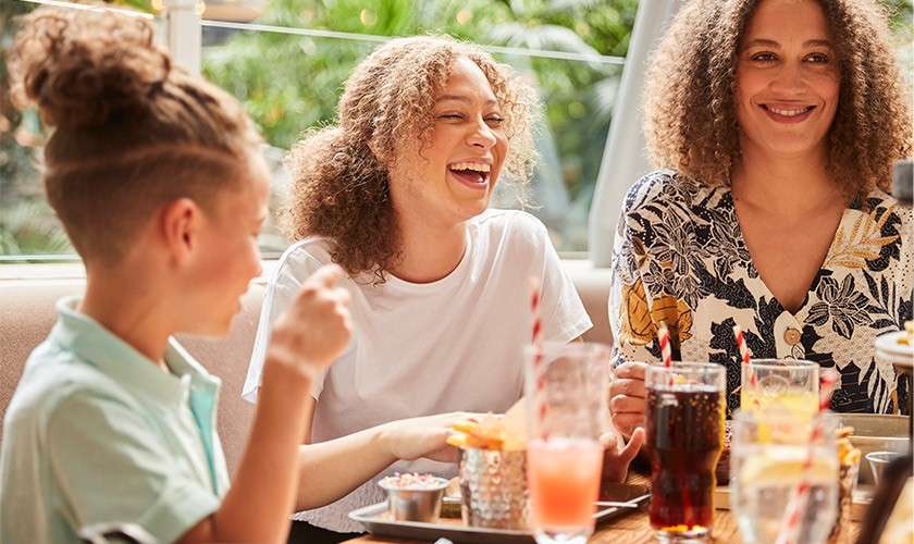 Three people laugh while dining. They sip drinks with striped straws and share fries on a wooden table, seated in a bright restaurant with big windows and leafy greenery outside.