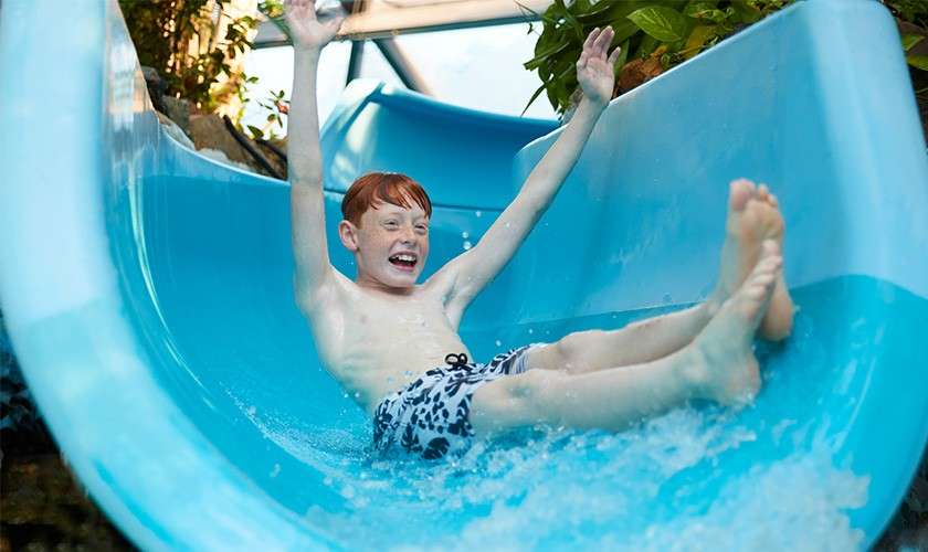 Child slides feet-first, arms raised, smiling, down a bright-blue water slide, splashing water. Context: indoor water park with sunlight through glass and surrounding plants along the slide.