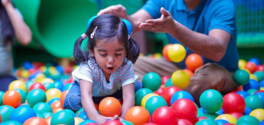 Child with pigtails reaches for plastic balls, mid-motion, mouth open in excitement; adult nearby tosses balls. Scene occurs in a colorful indoor ball pit with slide and netted play area.