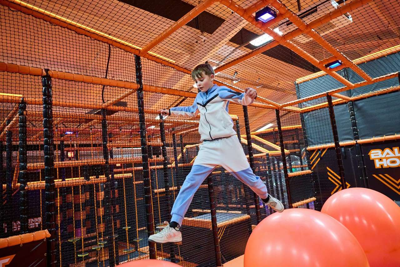 Child leaps between large inflated red balls, arms outstretched, in a netted indoor obstacle course with orange frames and padded walls. Visible text: BAL HO.
