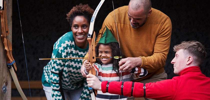 Child archer draws a bow with an arrow, smiling, while three adults guide and support hand and stance; they gather closely at an archery range, wearing sweaters and outdoor gear.