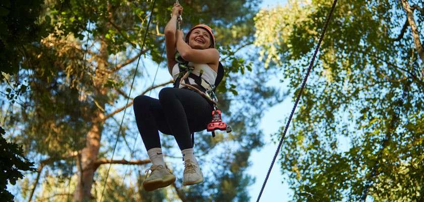 Person wearing a helmet and harness zips along a rope, legs bent and smiling, in a sunlit forest canopy with green leaves and tall trees surrounding.