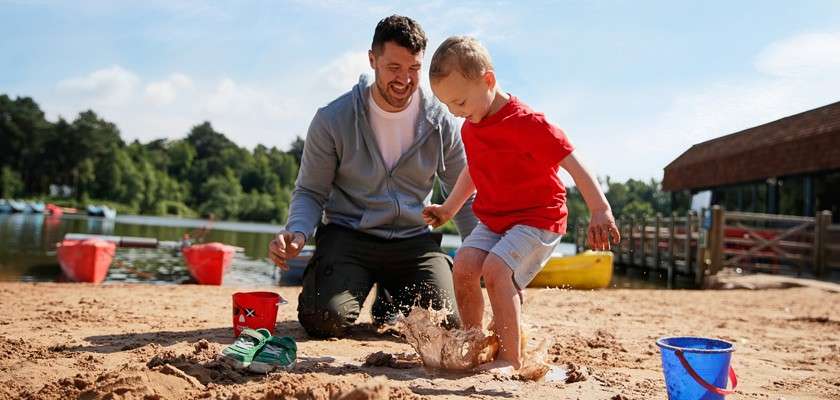 Child jumps and splashes muddy water with bare feet, as an adult kneels nearby. On a sandy lakeshore: buckets and sandals, calm lake, dock, trees, and colorful boats under sun.