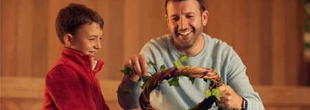 Wreath—being decorated with greenery by a smiling adult and child—indoors, under warm lighting, with a wooden backdrop and casual clothing.