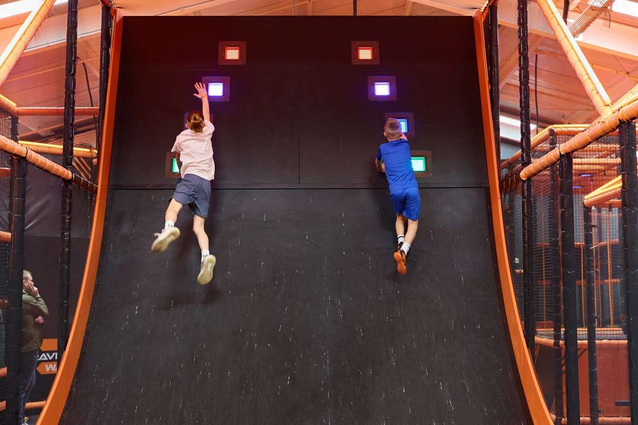 Two children run up a steep black wall, jumping to tap glowing colored buttons. They compete side-by-side inside an indoor adventure park framed by orange rails, netting, and overhead beams.