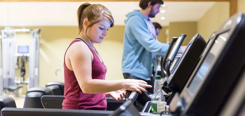 Person adjusts treadmill controls; in background, others walk on treadmills. Bright indoor fitness center with multiple machines, water bottle on console, soft lighting, beige walls.