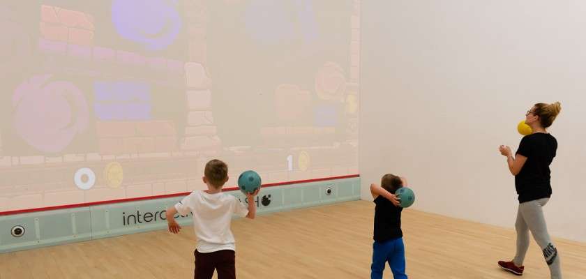 Three people prepare to throw soft balls at a projected game; two children and an adult face the screen in a bright indoor court. Text: interc, 0, 1.