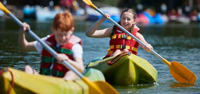 Two children in yellow kayaks paddle with double-bladed oars, wearing red life jackets, smiling. They move across a calm lake, with other colorful boats and trees blurred in the background.