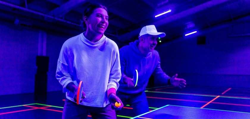 Two people play table tennis, laughing while holding paddles and a ball, under purple-blacklight illumination on a neon-lined court inside an indoor gym.