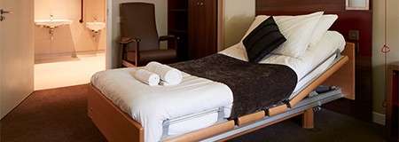 Adjustable bed sits inclined with pillows and rolled towels, placed in a tidy bedroom; chair, wardrobe, and open doorway reveal adjoining bathroom with twin sinks and warm lighting.