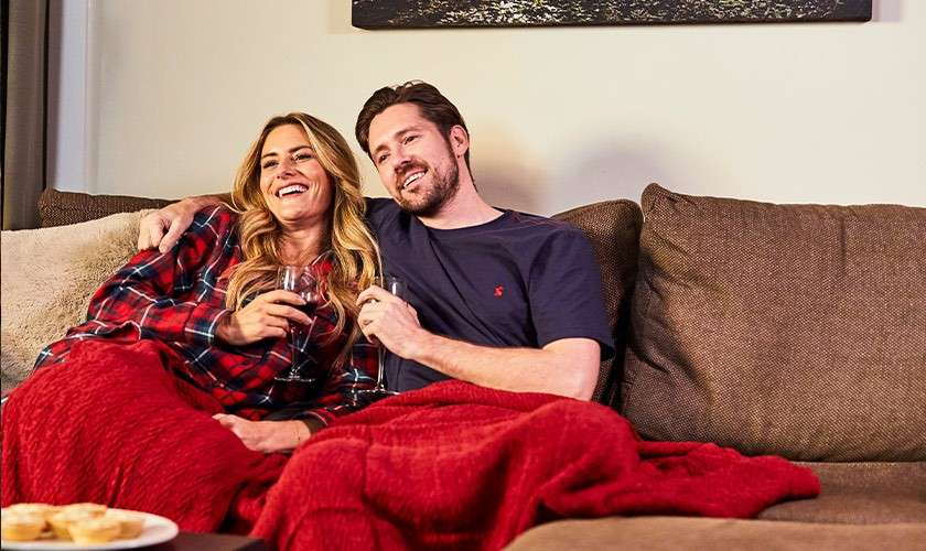 Couple relaxes, smiling and holding wine glasses, snuggled together under a red blanket. They sit on a brown sectional sofa in a cozy living room with soft lighting.