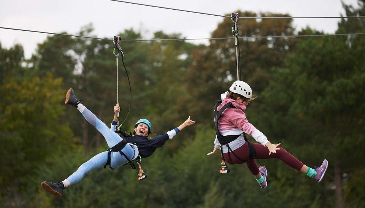 Two helmeted riders glide side-by-side on parallel zip lines, legs extended and arms raised, suspended above a forested area with tall green trees in soft light.